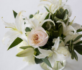 White lilies, white roses, and greenery in a glass vase on white surface—classic Holy Name of Jesus wedding bouquet.