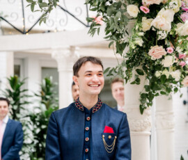 Groom smiles brightly as he stands under a floral mandap decorated with roses and greenery. He wears a navy blue embroidered sherwani with gold accents and a red pocket square, awaiting the bride during the wedding ceremony