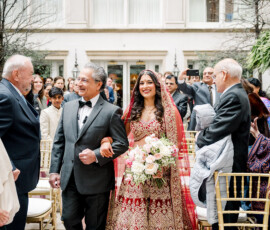 An Indian bride in a red and gold embroidered lehenga walks down the aisle with her father during an outdoor wedding ceremony. She carries a pastel bouquet and smiles as guests watch and take photos from both sides of the aisle.