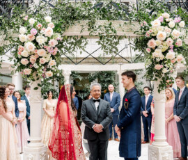 Bride and groom stand facing each other under an ornate floral mandap during their outdoor Indian wedding ceremony. The bride wears a red and gold embroidered lehenga with a matching veil, while the groom wears a navy blue sherwani. The officiant stands between them as their wedding party, dressed in coordinating pastel and navy attire, looks on.