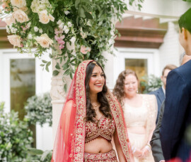 An Indian bride wearing a red and gold embroidered lehenga and matching dupatta smiles during the wedding ceremony under a floral mandap filled with roses and greenery. She stands facing the groom with guests watching behind her