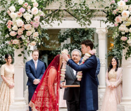 Groom places floral garland on bride at lively Indian wedding in New Orleans, with guests and floral decor in background.