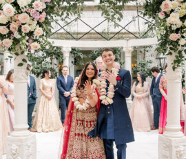 Smiling Indian couple shows rings under a floral arch at The Ritz-Carlton New Orleans, surrounded by their wedding party.
