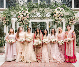 Bride dressed in a red and gold embroidered lehenga stands with her bridesmaids, who wear coordinated pastel lehengas in blush and champagne tones, each holding soft pink and white bouquets. They pose beneath a lush floral arch of roses and greenery in an elegant outdoor courtyard.
