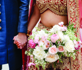 Close-up of a bride in a red and gold embroidered lehenga holding a lush bouquet of white and blush flowers with greenery. Her detailed blouse and dupatta shimmer with intricate beadwork, while she and the groom—dressed in a navy sherwani—hold hands just out of frame.