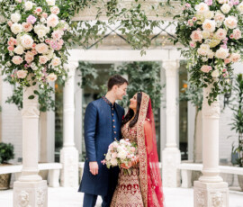Bride in red lehenga and groom in navy sherwani smile under floral arch at elegant Indian wedding, Ritz-Carlton New Orleans.