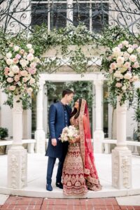 Bride in red lehenga and groom in blue sherwani smile under floral arch at New Orleans wedding ceremony.