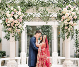 Bride and groom share an intimate kiss beneath a floral mandap adorned with roses and greenery. The bride wears a red and gold embroidered lehenga with a matching veil, while the groom wears a navy blue sherwani. They stand embraced under the ornate dome structure in the courtyard