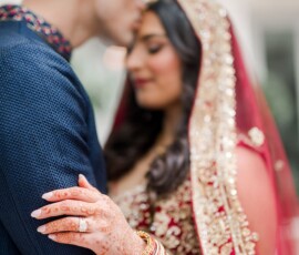 Groom kisses bride’s forehead as they embrace; she wears traditional attire, henna, bangles, and a red veil at their wedding.