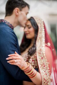 Groom kisses bride’s forehead as they embrace; she wears traditional attire, henna, bangles, and a red veil at their wedding.