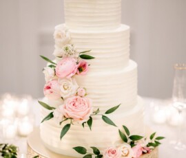 Elegant three-tier white wedding cake with pink and white flowers, green leaves, on a gold stand with votive candles.
