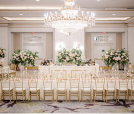 Grand wedding reception ballroom featuring a long tablescape with gold chiavari chairs, tall floral centerpieces of pink and white roses with greenery, and rows of floating candles. A large crystal chandelier hangs above, and a white bar with lush floral arrangements stands at the back of the room