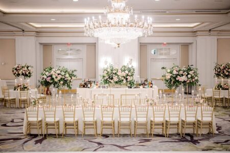 Banquet hall with gold chairs, white tables, pink and white floral centerpieces, crystal chandelier, and stylish bar in back.