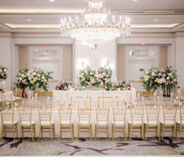Banquet hall with gold chairs, white tables, pink and white floral centerpieces, crystal chandelier, and stylish bar in back.