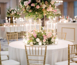 Round wedding reception table with gold chiavari chairs, featuring a tall floral centerpiece of pink and white roses with lush greenery. Clear cylinder candles surround the base of the arrangement, creating a romantic, glowing tablescape under the ballroom chandeliers.