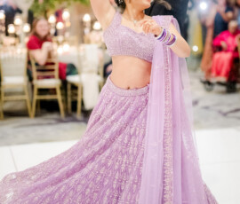 Bride twirls on the dance floor in a sparkling lavender lehenga as her groom spins her during their reception. She smiles brightly, her bangles and dupatta flowing with the movement while guests watch in the softly lit ballroom