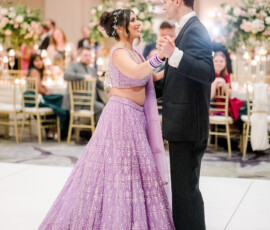 Bride and groom share their first dance at the reception. The bride wears a sparkling lavender lehenga with matching bangles and a jeweled hairpiece, smiling at her groom in a black tuxedo as guests watch in the background