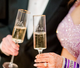 Close-up of the bride and groom holding personalized champagne flutes labeled ‘Mr.’ and ‘Mrs.’ during their wedding reception. The bride’s henna-covered hand, purple bangles, and engagement ring are in focus beside her sparkling lavender lehenga