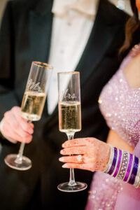 A couple toasts with champagne at their wedding; her glass reads Mrs., showing henna and purple bangles for unique inspiration.