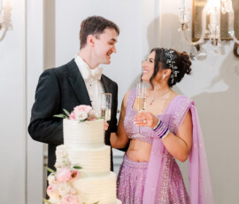 Couple in tuxedo and lavender sari smile by a white cake with pink flowers, celebrating at a romantic New Orleans wedding.