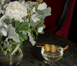 Martini with two olives on dark marble table, white flowers, and red velvet chair at The Columns Hotel in New Orleans.
