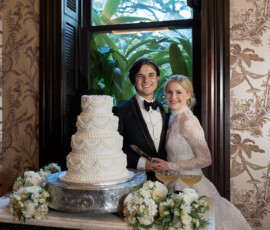 Bride and groom smile by a tall white wedding cake at The Columns Hotel in New Orleans, with florals and greenery visible.