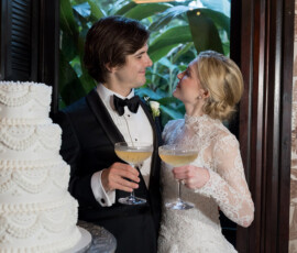 Bride and groom smiling with champagne by a white wedding cake at The Columns Hotel, New Orleans.