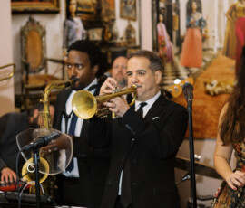 Two suited musicians play trumpet and saxophone amid elegant decor at a New Orleans-style wedding, with bandmates in the background.