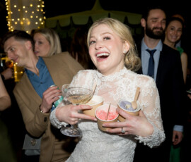 Bride in white lace dress smiles, holding champagne and gelato at lively New Orleans wedding with dancing guests.
