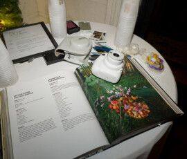 White instant camera on cookbook with salad photo, cups, pens, photos, and framed menu on round white table at New Orleans wedding.