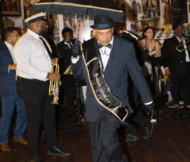 Man in suit and sash dances with black umbrella, leading a New Orleans wedding band at The Columns Hotel.