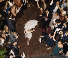 Crowd at outdoor New Orleans wedding, guests waving handkerchiefs as a person in white walks with a decorative umbrella.