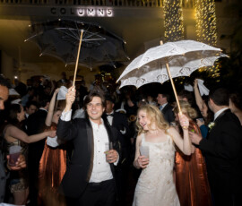 A smiling couple in formal wear hold decorated umbrellas at a lively New Orleans wedding outside The Columns Hotel at night.