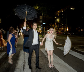 Formally dressed couple with umbrellas smile, walk hand in hand at night amid joyful New Orleans wedding celebration.