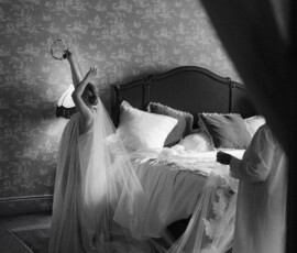 Bride in flowing gown holds flower crown by bed; another stands nearby, Columns Hotel New Orleans wedding room glows softly.