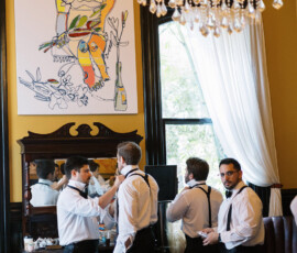 Four men in formalwear adjust bow ties and suspenders in a bright room at The Columns Hotel before a New Orleans wedding.