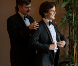 Two men in tuxedos smile as they prepare for a New Orleans wedding at The Columns Hotel, sunlight and greenery nearby.