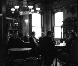 Men in suits sit at tables in a vintage, dim bar with large windows at The Columns Hotel, New Orleans wedding.