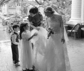Bride in lace gown on The Columns Hotel porch, aided by two kids and a woman; outdoor setting with trees and houses.