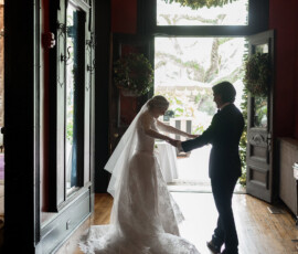Bride and groom hold hands in a sunlit doorway, framed by greenery at their intimate Holy Name of Jesus wedding.