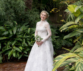 Bride in long-sleeved lace dress smiles with bouquet on brick path amid lush tropical plants at Holy Name of Jesus, New Orleans.