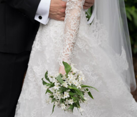 Bride in lace dress with white bouquet stands next to groom in dark suit, lush greenery at The Columns Hotel, New Orleans.