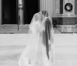 Two brides in wedding dresses and veils kiss outside a church in this black-and-white, elegant wedding photo.