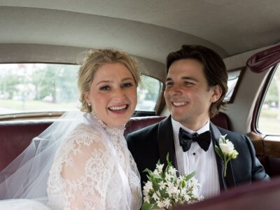 Bride in lace dress and groom in tuxedo smile together with white flowers in a vintage car on their Louisiana wedding day.