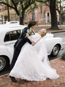 A groom dips his smiling bride in a white dress by a classic car on a tree-lined street—elegant Louisiana wedding inspiration.