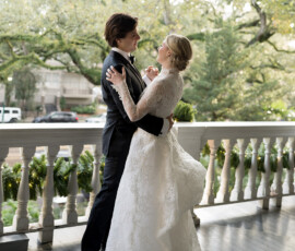 Bride and groom dance on a greenery-decorated porch at The Columns Hotel in New Orleans; trees and cars in background.