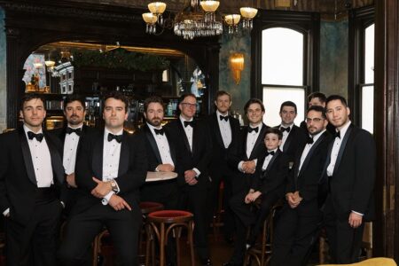 Thirteen males in tuxedos pose in a vintage bar with chandeliers before a Louisiana wedding ceremony.