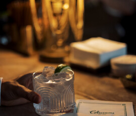 Hand holds a clear cocktail with ice and lime at The Columns Hotel bar; napkins, lamp, and MEET ME AT THE COLUMNS coaster nearby.