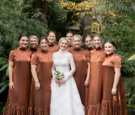 Bride in white lace gown with eight bridesmaids in rust dresses amid greenery at The Columns Hotel, New Orleans wedding.