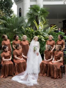 Bride in white dress with bouquet and eight bridesmaids in copper satin outside New Orleans wedding, greenery and white building.